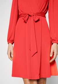 Červené šaty s dlouhými rukávy a elastickými manžetami, opasek na zavázání a hladký, splývavý materiál. Spodní lem je mírně asymetrický.