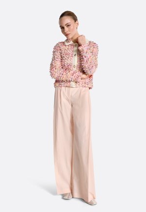 Ženska, oblečena v teksturirano rožnato-kremasto jakno z gumbi, širokimi bledo rožnatimi hlačami in kremastimi vzorčastimi čevlji, stoji z eno roko prekrižano.