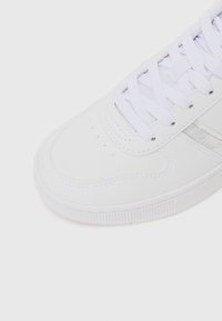 Hvit sneaker med en glatt overdel, rund tå og snøring. Merkbar sølvdetalj på siden og teksturert gummisåle.
