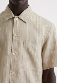 Camicia a maniche corte con bottoni di colore beige chiaro con sottili righe bianche, realizzata in tessuto testurizzato, dotata di una tasca sul petto e bottoni in legno.