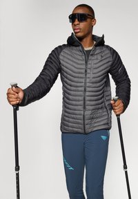 Veste noire rembourrée avec capuche et design matelassé, associée à un pantalon bleu marine. L'individu tient des bâtons de randonnée et porte des lunettes sombres.