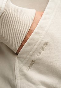 Χέρι στην τσέπη ενός ανοιχτό μπεζ φούτερ με ριμπ μανσέτα και διακριτικό κεντημένο λογότυπο "SANWOOD" κοντά στη ραφή της τσέπης.