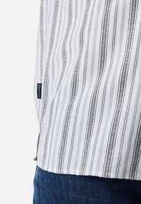 Gestreept shirt van wit en zwart stof, met verticale lijnen en een zijkantlabel. De textuur lijkt licht en ademend te zijn.