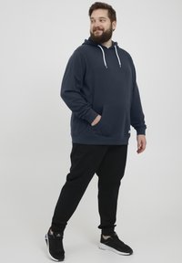 Sudadera con capucha azul marino y un bolsillo delantero, cordones blancos y pantalones jogger negros. Zapatillas deportivas negras completan el conjunto.