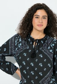 Blusa negra con un estampado floral en azul claro, mangas largas, escote redondo y un detalle de lazo adornado con una borla negra.