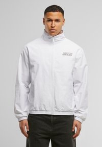 White zip-up jacket with high collar, elastic cuffs, and hem. Features a logo on the chest. Made of smooth, lightweight fabric.