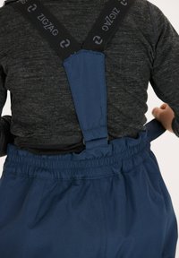 Salopette bleu marine avec taille élastique et bretelles noires ajustables surmontées d'un logo en zigzag. Le matériau semble durable et texturé.
