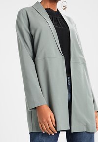 Cardigan lungo verde chiaro a maniche lunghe con collo a scialle, caratterizzato da una vestibilità rilassata e da una texture liscia, indossato sopra una maglia nera e jeans di denim.