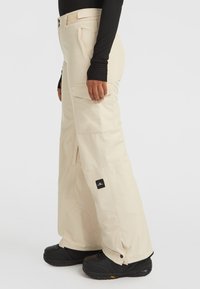 Beige sneeuwbroek met een relaxed fit, voorzien van grote cargozakken, verstelbare manchetten en een logo patch, gecombineerd met zwarte sneeuwschoenen.