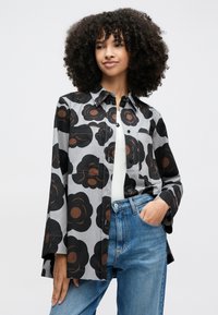 Young woman with curly hair wearing a gray button-up shirt with large black and brown floral patterns, white top, and blue jeans.