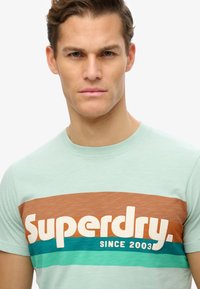 Mint green t-shirt featuring bold "Superdry" logo in white, with horizontal stripes in brown and teal across the chest. Smooth texture.