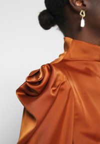 Satinbluse in verbranntem Orange mit sanftem Glanz, ausgestattet mit einer drapierten Schulterpartie und einem hohen Kragen. Kleine runde Ohrringe mit Perlenakzent.