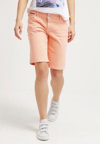 Shorts de mezclilla color durazno, con corte recto, bolsillos frontales y un solo botón. Combinados con zapatillas grises de velcro.