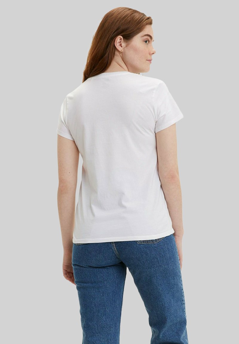 Frau mit langen braunen Haaren, die ein schlichtes weißes kurzärmeliges T-Shirt und blaue Jeans trägt, steht mit dem Rücken zur Kamera vor einem neutralen Hintergrund.
