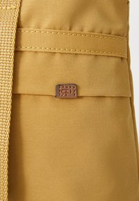 Yellow fabric backpack with textured material, featuring a sewn leather label and woven strap detail. Compact design with a front pocket.