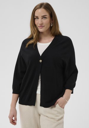 Kobieta o jasnobrązowych włosach, ubrana w czarny kardigan na guziki na białą koszulę i beżowe spodnie, z jedną ręką w kieszeni.