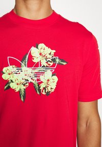 Osoba v červeném tričku s bílým obrysem loga Adidas, které se nachází přes žlutý a zelený květinový vzor na hrudi.