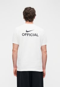 Balti medvilniniai marškinėliai su juodu Nike logotipu ir ant nugaros atspausdintu užrašu „OFFICIAL“, trumpomis rankovėmis ir apvalia iškirpte.