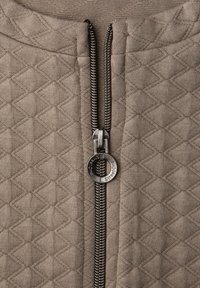 Beige Reißverschlussjacke mit gestepptem Diamantmuster, ausgestattet mit einem Metallreißverschluss und rundem Zugtab, gefertigt aus weich texturiertem Stoff.