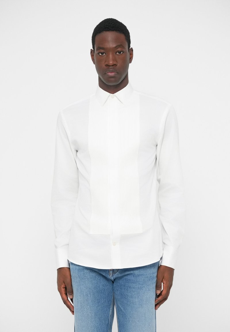 Λευκό πουκάμισο μακρύ μανίκι με πτυχωτό μπροστινό πάνελ, κλασικό γιακά και κουμπί κλεισίματος. Φοριέται με ανοιχτό μπλε τζιν παντελόνι.