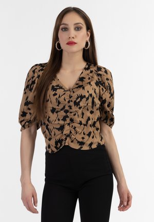 Vrouw met lang bruin haar met parel-oorbellen, een bruin-zwarte blouse met geplooide details en zwarte broek tegen een witte achtergrond.