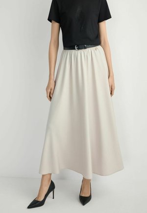 Kobieta ubrana w czarny t-shirt, beżową spódnicę do kostek z czarnym paskiem oraz czarne szpilki w szpic, stojąca na jednolitym tle.