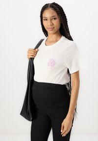 T-shirt en coton blanc avec un design floral rose sur la poitrine, accompagné de pantalons noirs extensibles. Le modèle tient un sac noir.
