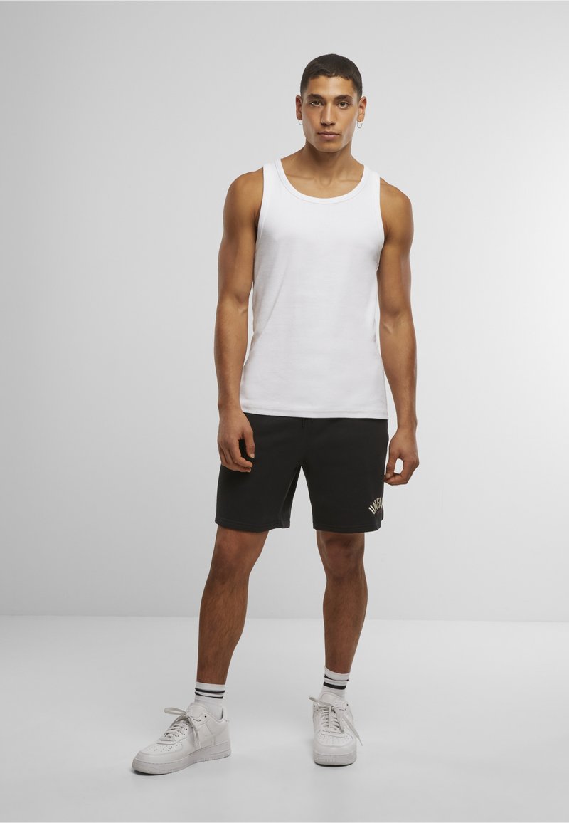 Mann trägt ein weißes Tanktop, schwarze Shorts mit Schriftzug, weiße Turnschuhe und gestreifte Socken, steht vor einem schlichten grauen Hintergrund.