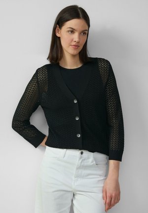 Zwarte gebreide cardigan met V-hals, driekwart mouwen, knoopsluiting en zigzagpatroon. Draagt over een zwarte top en witte broek.