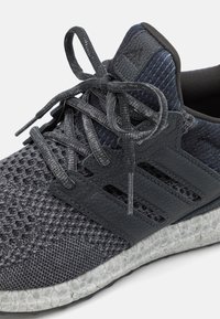 Athletic shoe with a mesh upper in shades of gray, textured overlays, and a white patterned outsole. Features lace-up closure and branding.