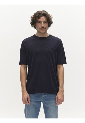 Man met krullend bruin haar en snor, gekleed in een donker marineblauw T-shirt en lichtblauwe spijkerbroek tegen een effen witte achtergrond.