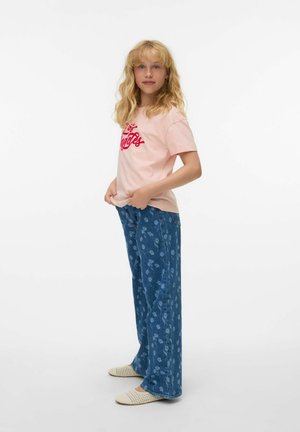Jeune femme blonde portant un t-shirt rose Los Angeles, un jean large à fleurs bleu, et des chaussons tissés beige, debout devant un fond blanc.
