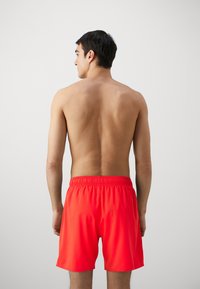 Spilgti sarkani peldbikses ar elastīgu jostu, gluda tekstūra, līdz augšstilbiem, vienkāršs dizains un bez redzamām drukām.