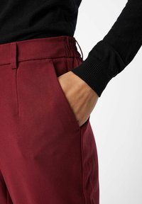 Un pantalon bordeaux présente un design épuré avec une ceinture élastique, des poches latérales et une texture douce en coton. Un manchon noir côtelé est visible.