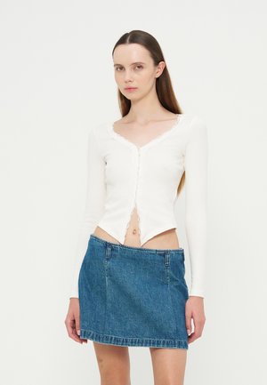 Jeune femme portant un haut blanc à manches longues et une jupe en jean bleue, debout devant un fond uni.