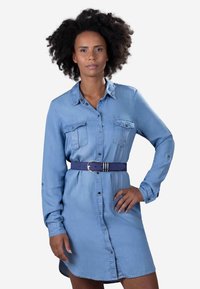 Robe chemise en denim bleu clair avec un devant à boutons, deux poches poitrine et une large ceinture bleu marine. Manches longues et coupe décontractée.