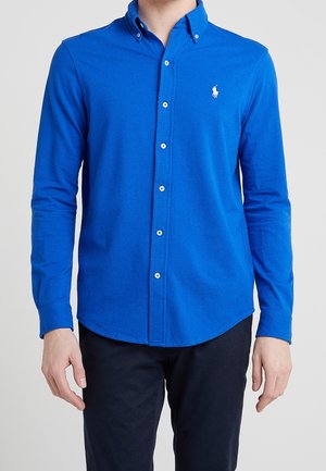 Homme portant une chemise ajustée à manches longues de couleur bleue vif, boutonnée, avec un petit logo blanc brodé sur la poitrine gauche.