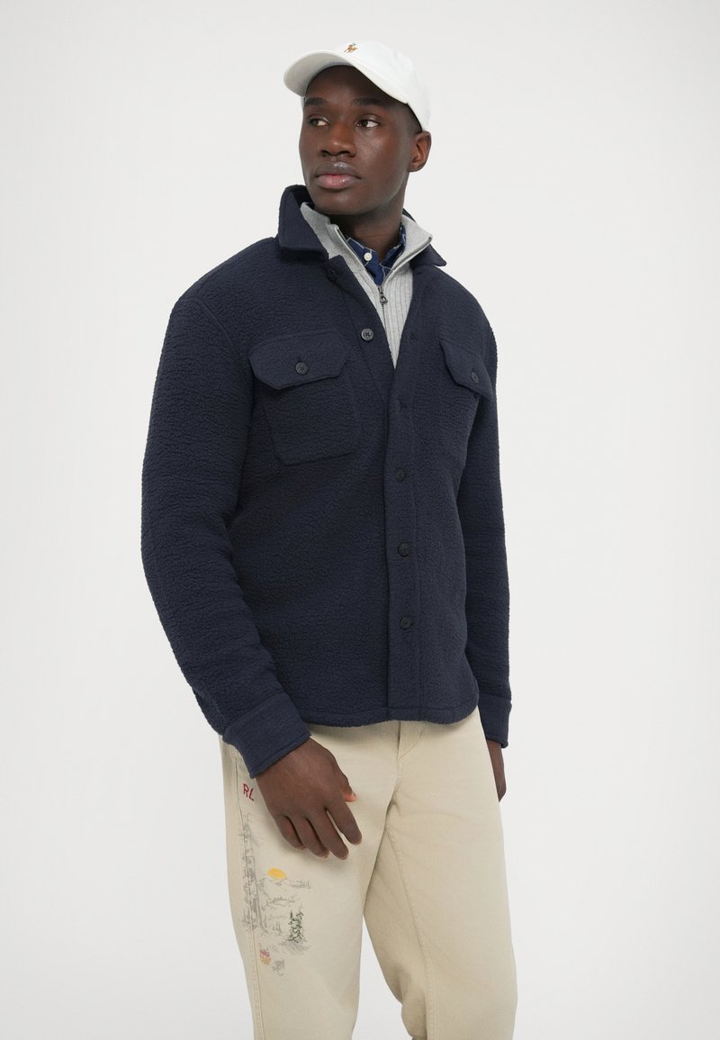 Navy fleece jacket with two chest pockets, button closure, and textured fabric. Paired with light beige pants featuring an embroidered design.