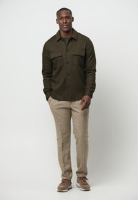 Camicia verde con bottoni e due tasche sul petto, pantaloni beige texturizzati e sneakers marroni. Design semplice, vestibilità rilassata e dettagli minimi.