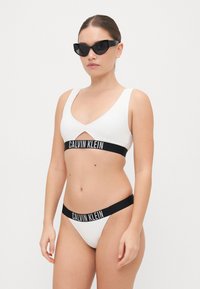 Bikini blanc à côtes avec une bande de logo Calvin Klein noire. Stylé avec des lunettes de soleil noires en forme de chat. Texture lisse et design épuré.