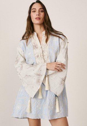 Mujer con un vestido de patrón azul claro y crema, de mangas anchas y con flecos, de pie con los brazos cruzados frente a un fondo liso.