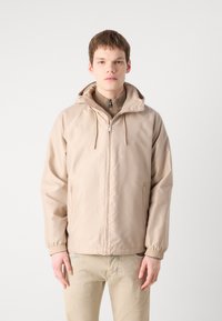 Beige nylonjacka med huva, framsida med dragkedja och elastiska ärmslut. Har sidofickor och en mjuk, lätt textur.