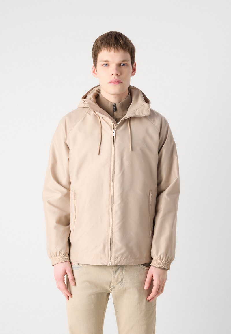Veste en nylon beige avec capuche, fermeture éclair à l'avant et poignets élastiques. Dispose de poches latérales et d'une texture lisse et légère.