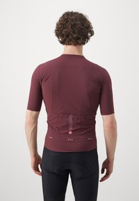 Bordeauxrotes kurzärmliges Fahrradtrikot mit strukturiertem Finish, ausgestattet mit einer Rückentasche, Logo und reflektierenden Akzenten. Kombiniert mit schwarzen Radhosen.