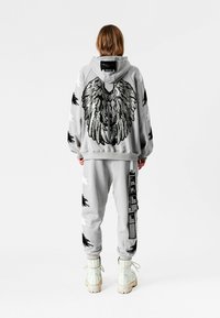 Persona con sudadera con capucha y pantalones grises con diseños de plumas en blanco y negro, patrón de alas plateadas en la espalda y botas blancas con cordones, de pie.