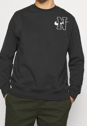 Mężczyzna w czarnej bluzie z białym "N" i logo swoosh na piersi oraz w ciemnozielonych spodniach, ręce w kieszeniach.