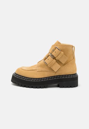 Zamszowe botki za kostkę w jasnym żółtym kolorze z czarną gumową podeszwą. Posiadają dwa poziome paski i srebrne elements. Szwy dekoracyjne wzdłuż podeszwy.