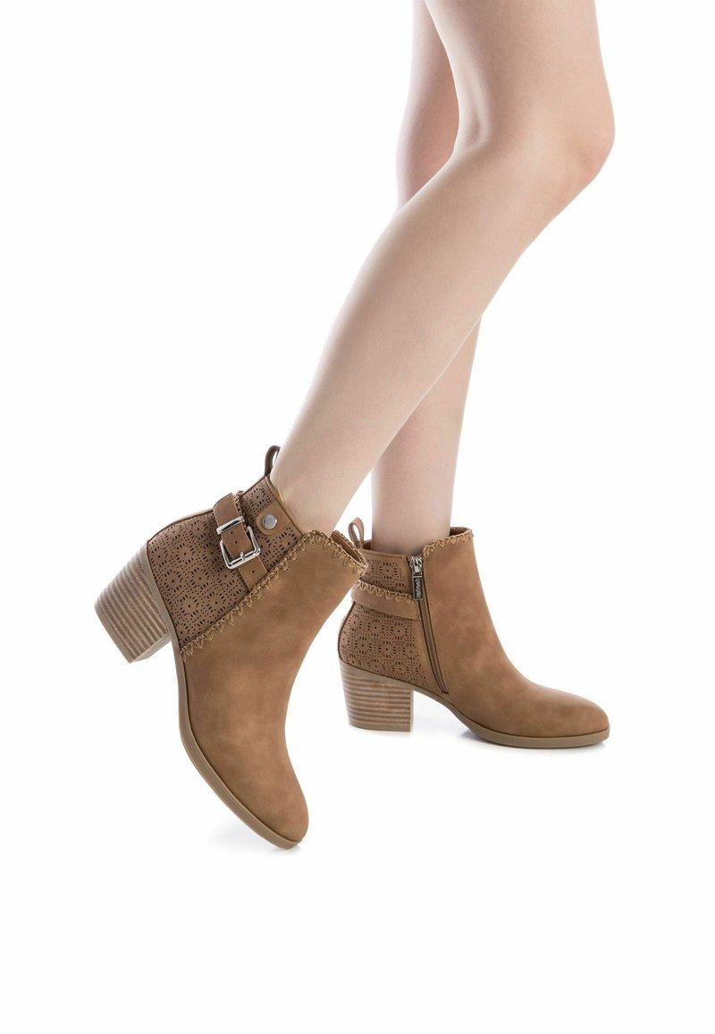 Stivaletti alla caviglia marroni in suede con un design perforato, zip laterale, fibbia regolabile e tacco a blocco. Texture liscia e punta arrotondata.