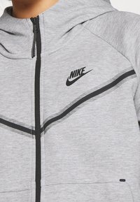 Grå zip-up huvtröja i mjukt tyg, med svarta detaljer, en iögonfallande Nike-logga och en zip-up fram med hög krage.