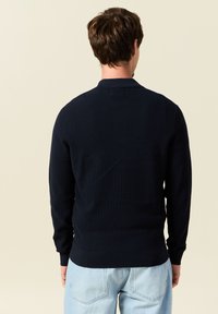 Tummansininen ribbikuvioinen pitkähihainen neulepaita, jossa on kaulus, tarjoillen vartalonmyötäistä muotoilua ja sileää tekstuuria, yhdistettynä vaaleansiniin farkkuihin.